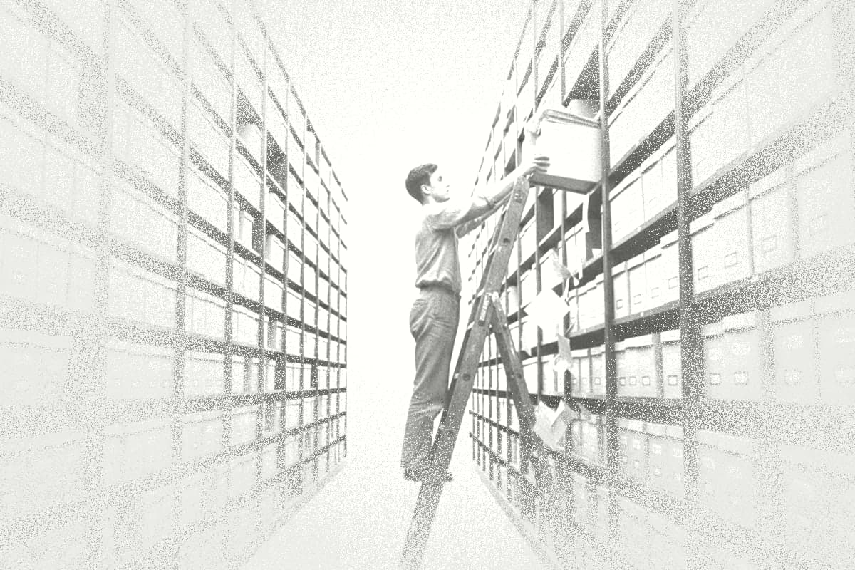 A young man on a ladder in an archive pulls a box of documents from tall shelves.