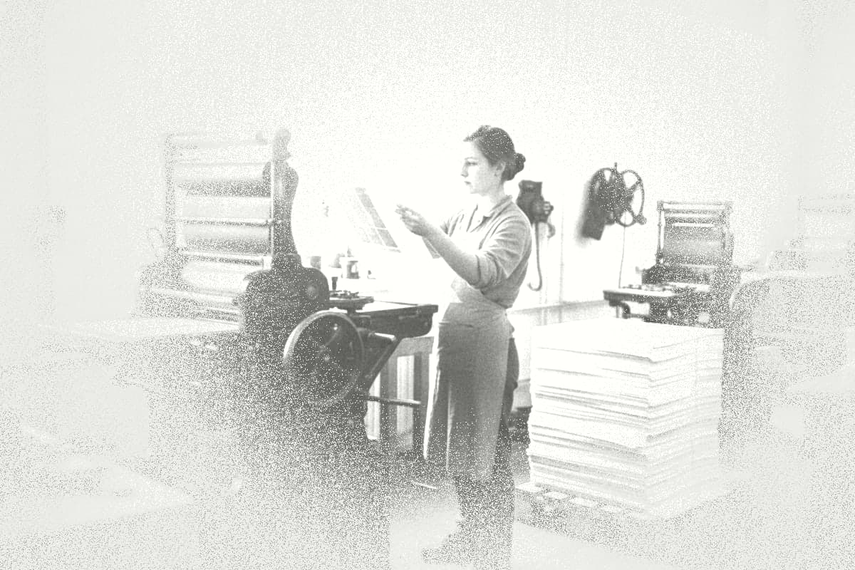 A young woman in a print shop holds a freshly printed sheet up to the light.